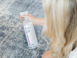 women with blonde hair holding allergen spray bottle in hand spraying on rug