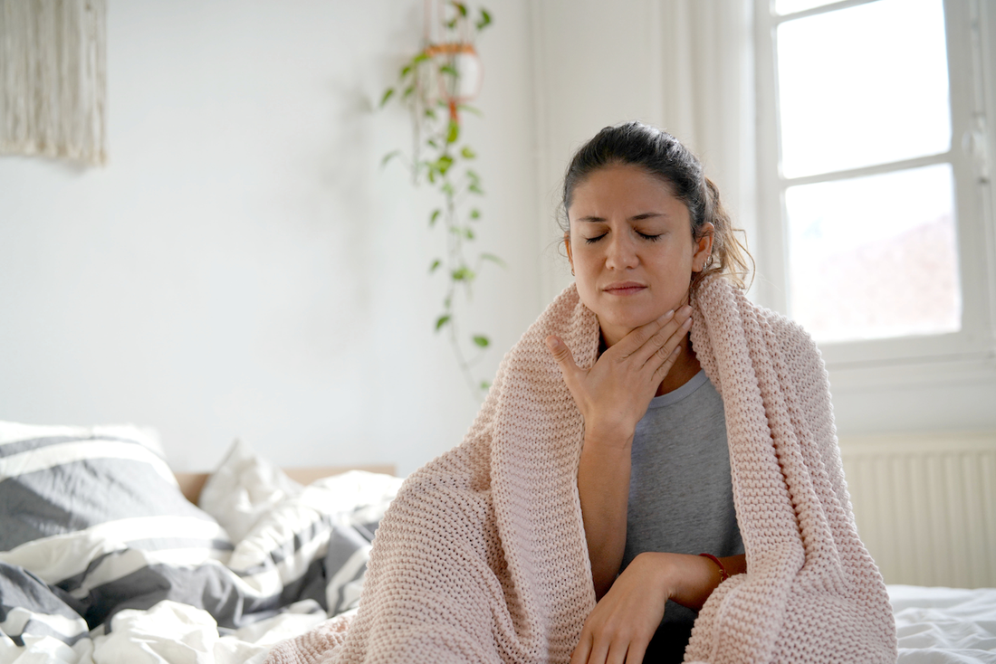 photo of a young women holding her throat looking sick while in her bedroom wrapped in a blanket