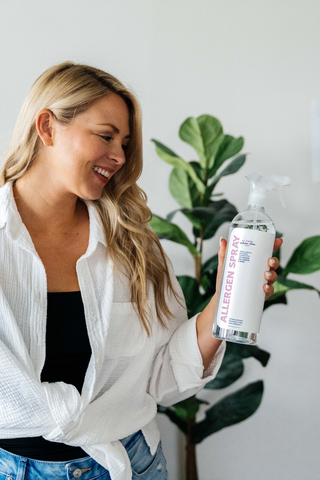 allergen spray by allergy defender bottle with a women holding bottle in front of plant on white background